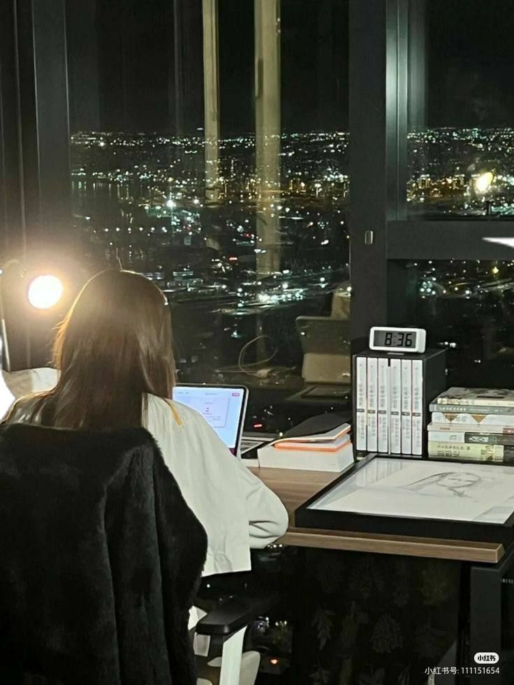 This may contain: a woman sitting at a desk in front of a window with a laptop computer on it