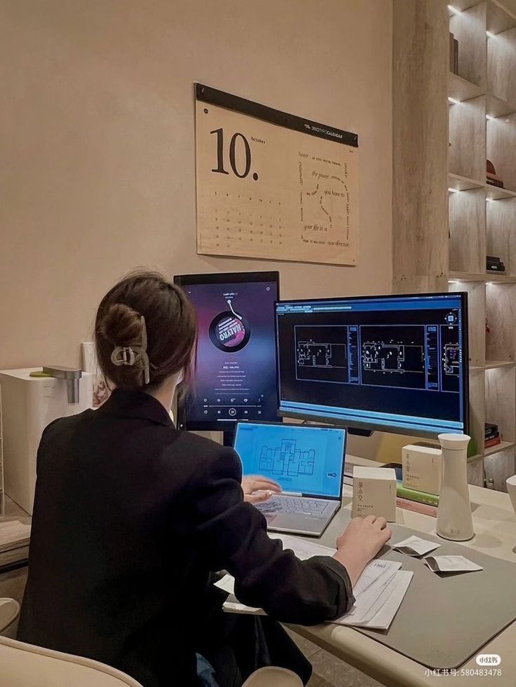 This may contain: a woman sitting at a desk with two laptops and a desktop computer on it