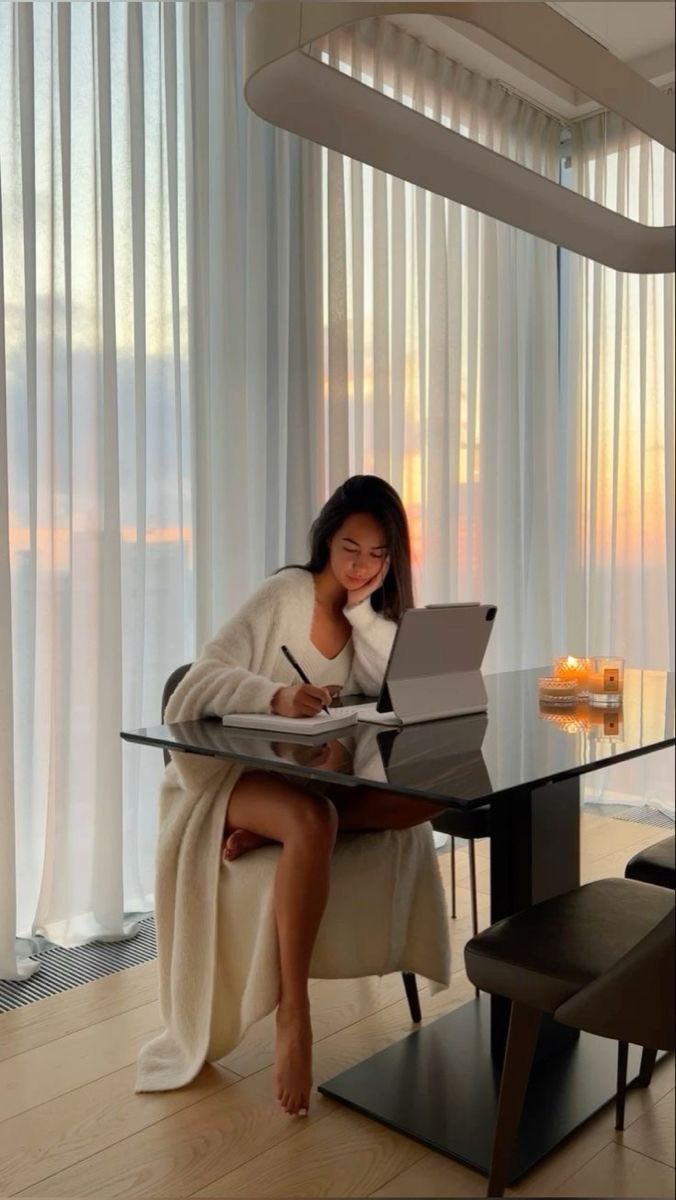 This may contain: a woman sitting at a table with a laptop computer in front of her and writing