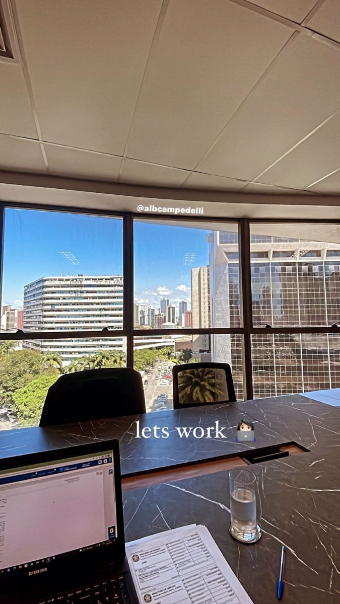 This may contain: an office desk with a laptop and papers on it in front of large windows overlooking the city