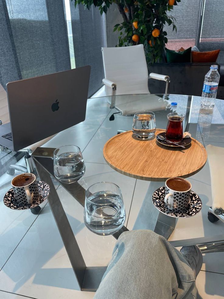 This may contain: a glass table topped with cups and saucers next to a laptop computer on top of a white tiled floor