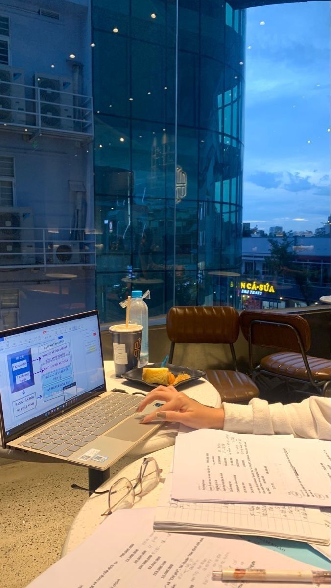 This may contain: a person sitting at a table with a laptop computer and papers on it, in front of a large window