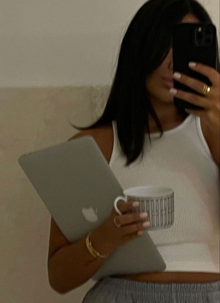 This may contain: a woman holding a laptop computer and looking at her cell phone while holding a cup