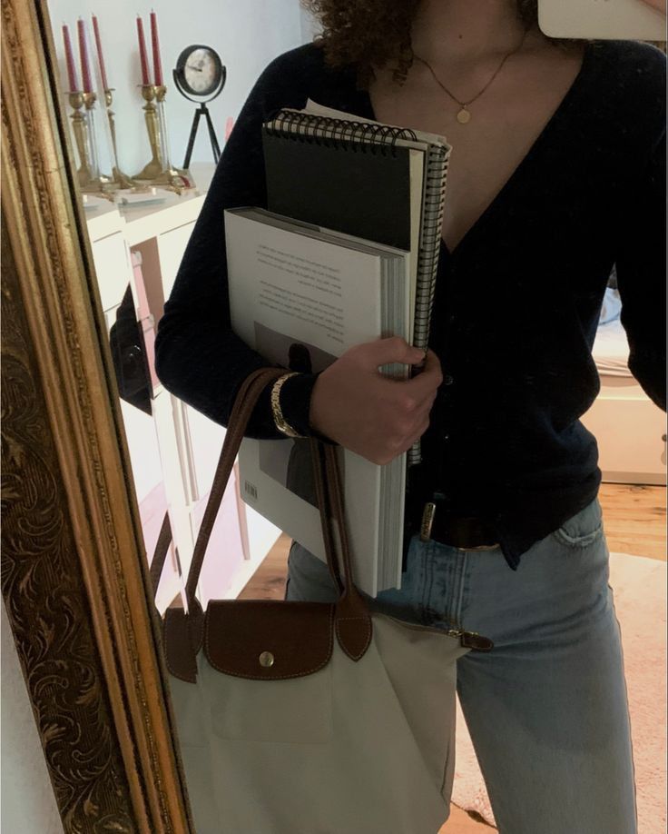 This may contain: a woman is holding a book in her hand and looking at the camera while standing next to a mirror