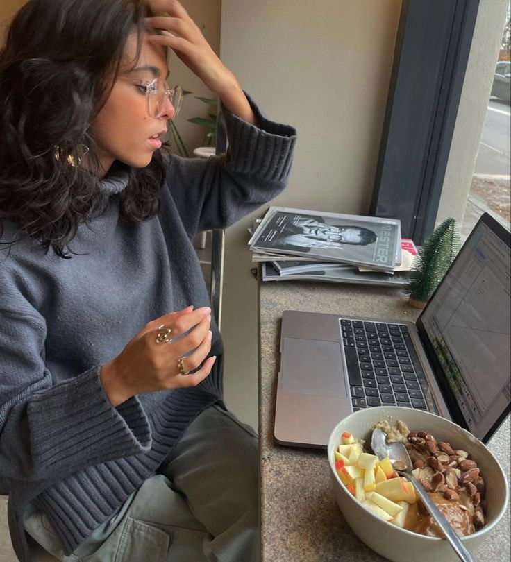 This may contain: a woman sitting at a table with a bowl of food in front of her laptop