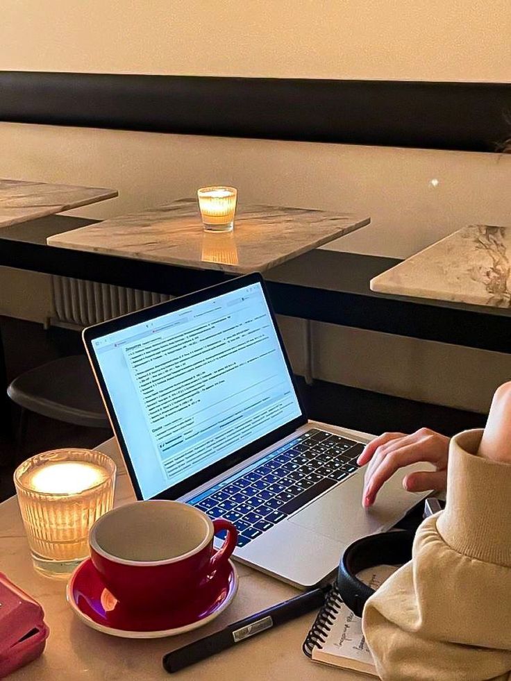This may contain: a woman sitting at a table with a laptop computer on her lap and coffee mug in front of her