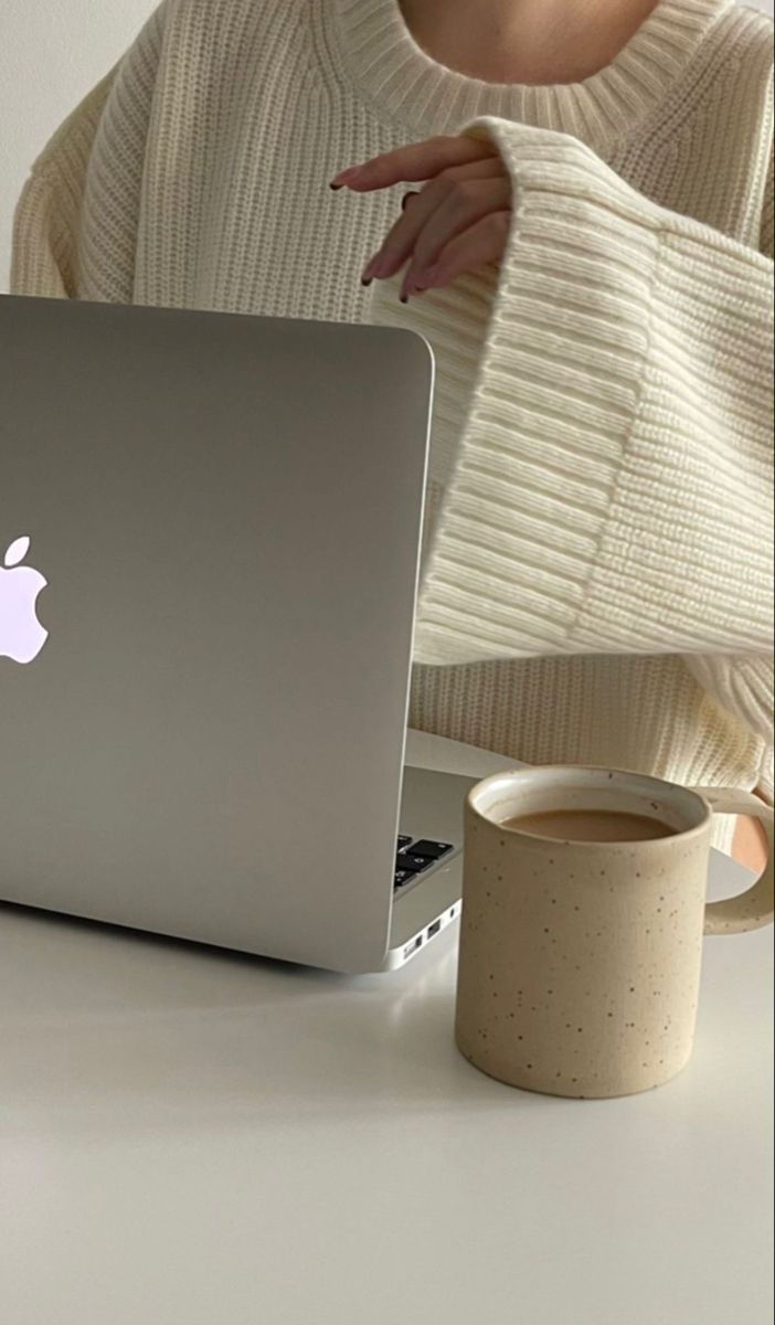 This may contain: a woman sitting at a table with an apple laptop in front of her and a cup of coffee next to her