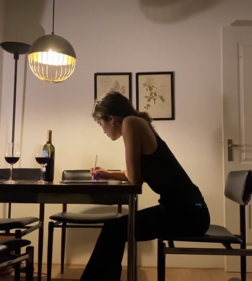 This may contain: a woman sitting at a table writing on a piece of paper with a bottle of wine in the background