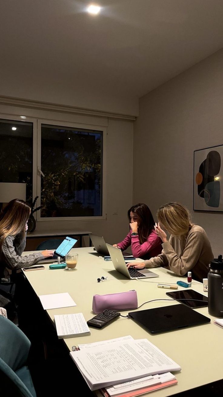 This may contain: three women sitting at a table with laptops and papers in front of their faces