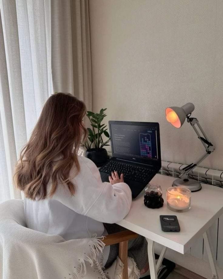 This may contain: a woman sitting in front of a laptop computer on top of a white desk next to a window