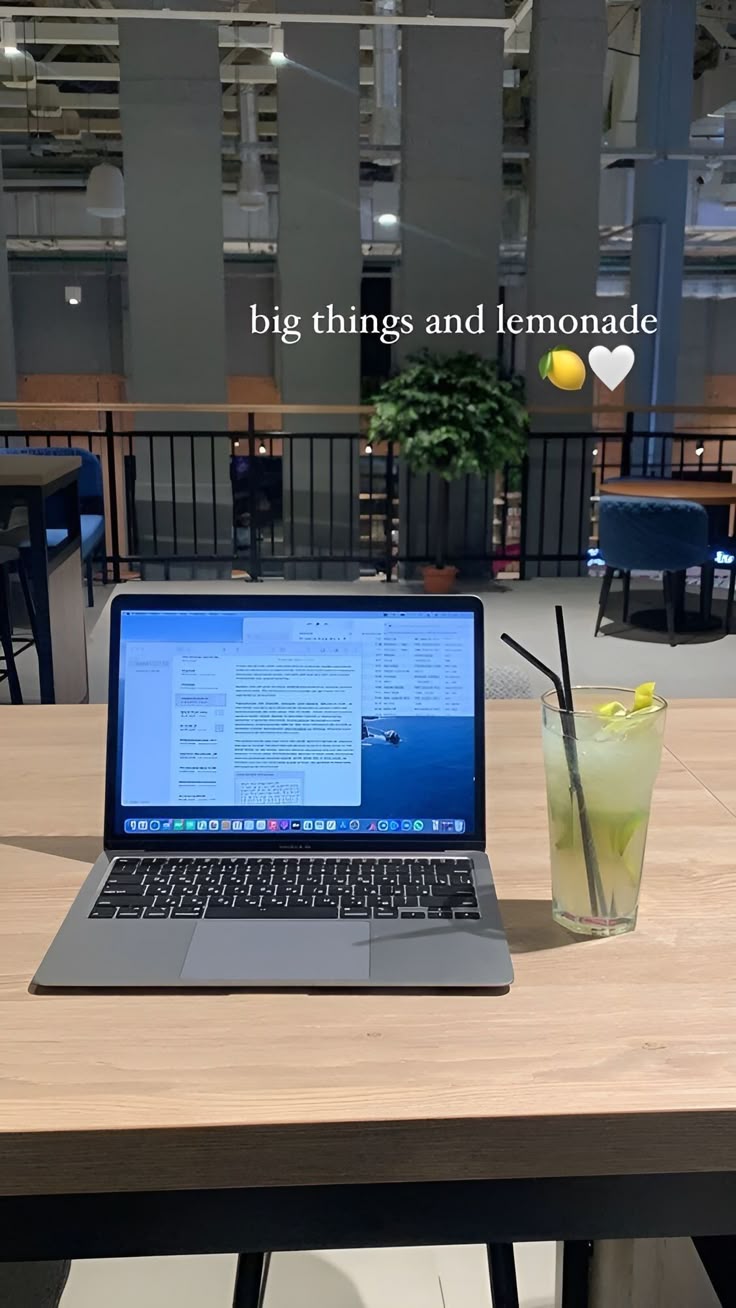 This may contain: an open laptop computer sitting on top of a wooden table next to a glass filled with lemonade