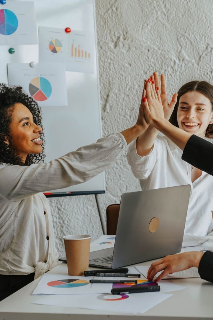 This may contain: three women giving each other high fives in front of a laptop on a table