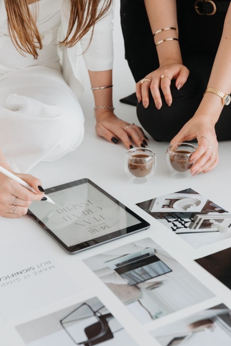 This may contain: two women sitting at a table with some pictures on the table and one holding a tablet
